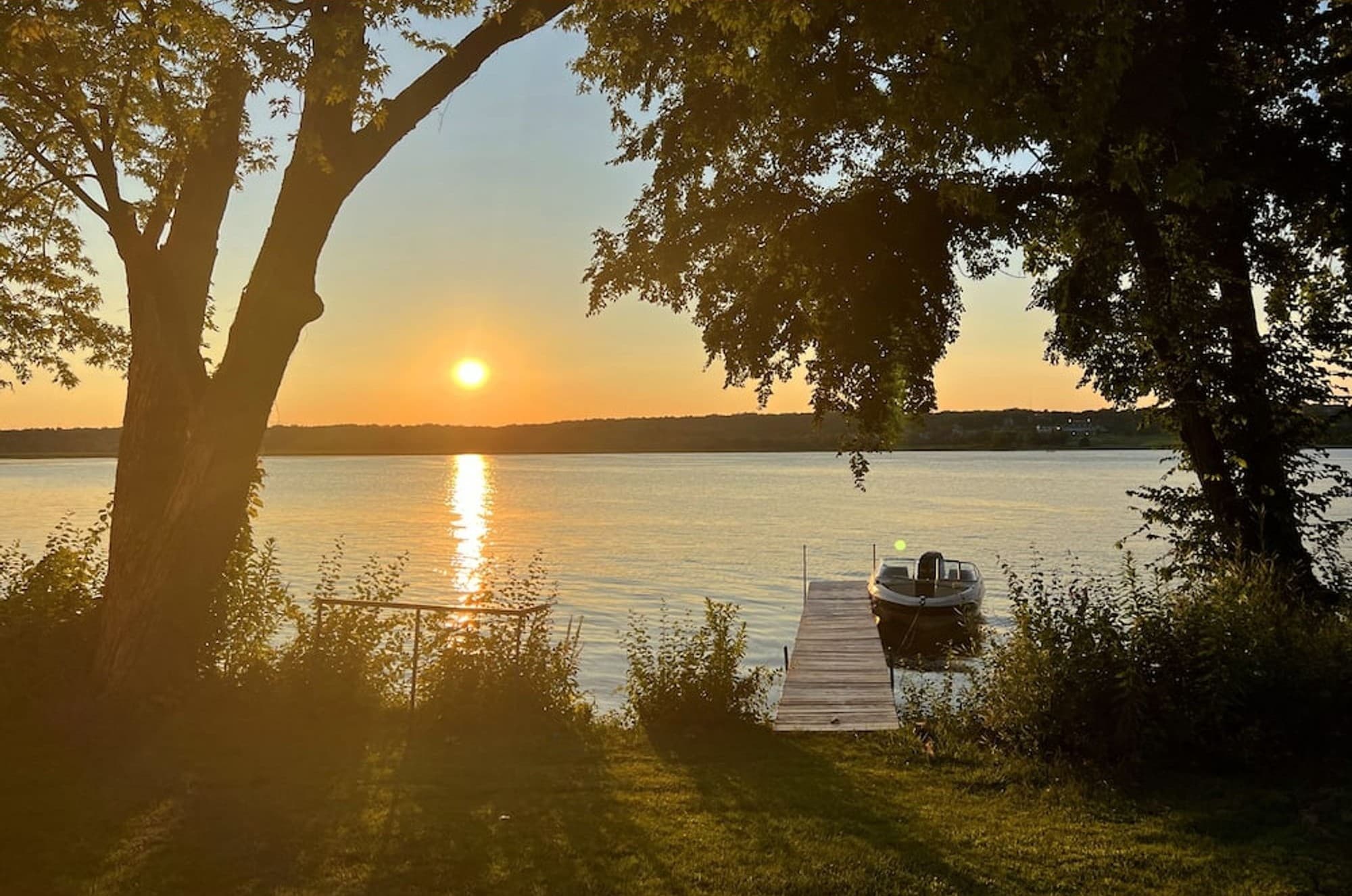 The sun setting over the lake, with the pier and a boat below the trees